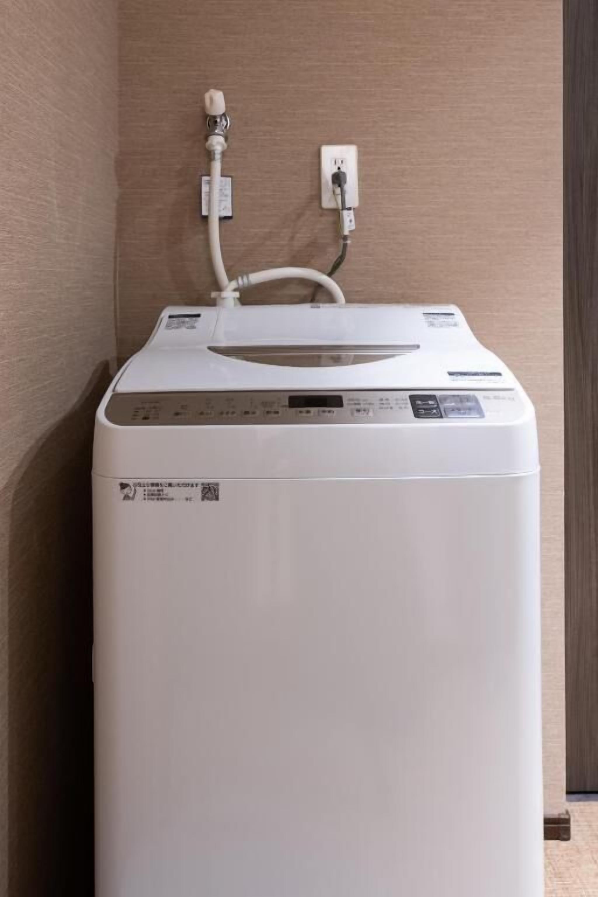 Eine kompakte, weiße, an der Wand montierte Waschmaschine mit Bedienfeld, einem Stromanschluss und einem Wasseranschluss an einer beige gefärbten Wand.