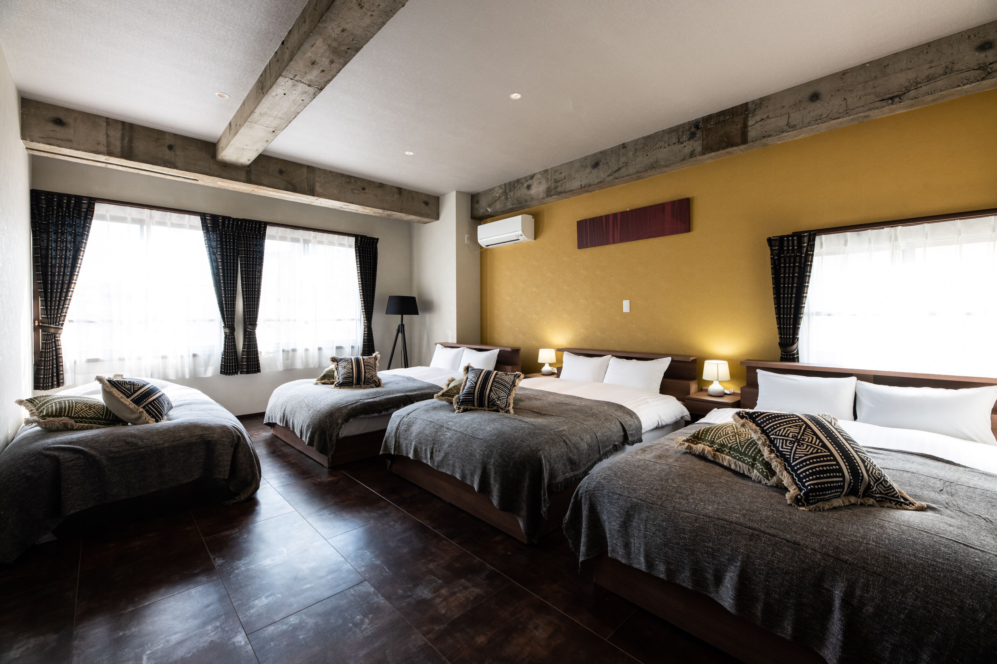 A hotel room with three beds, wooden flooring, a yellow accent wall, and large windows with curtains.