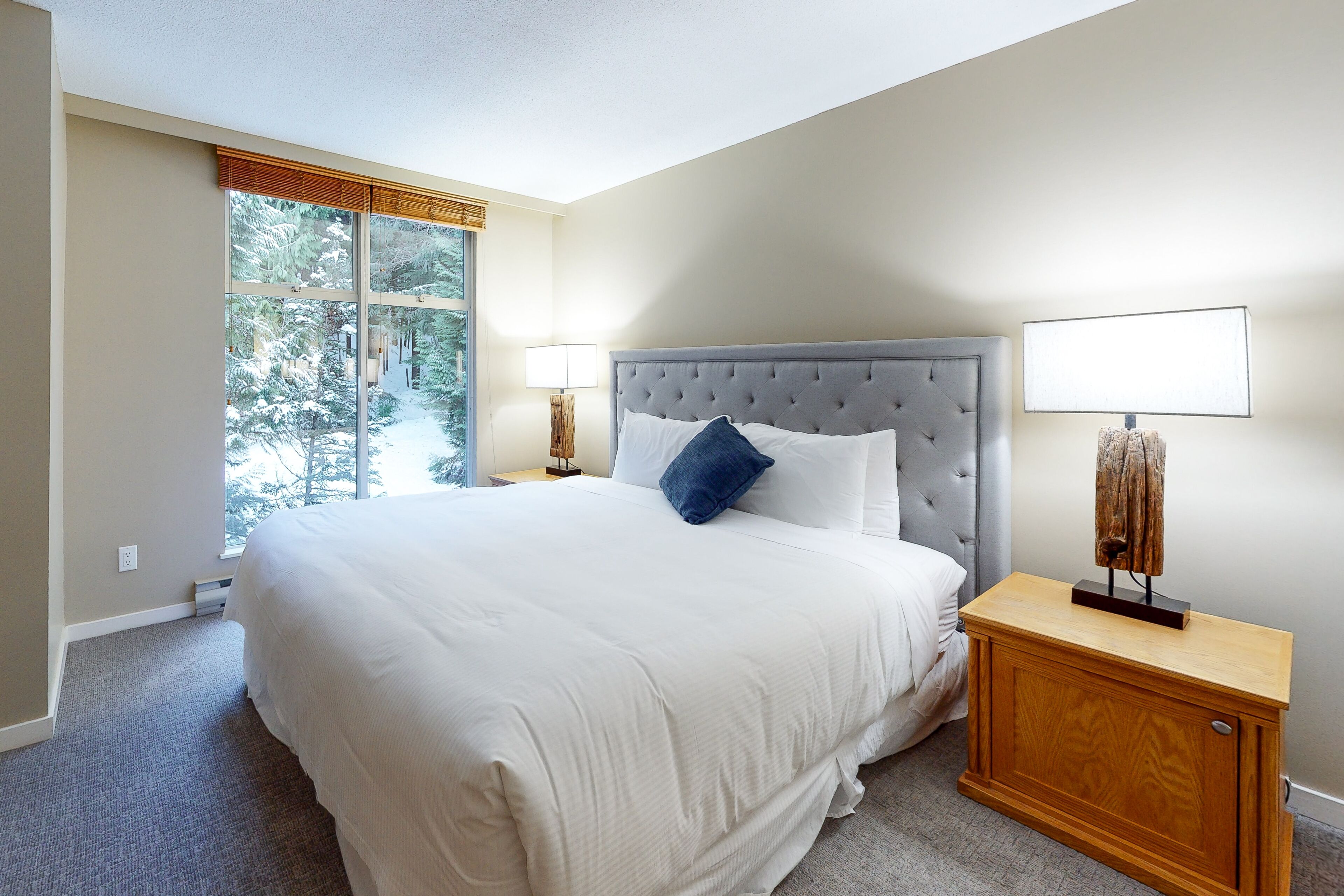 A bedroom with a large bed, two bedside lamps, a nightstand, and a window with a view of trees.