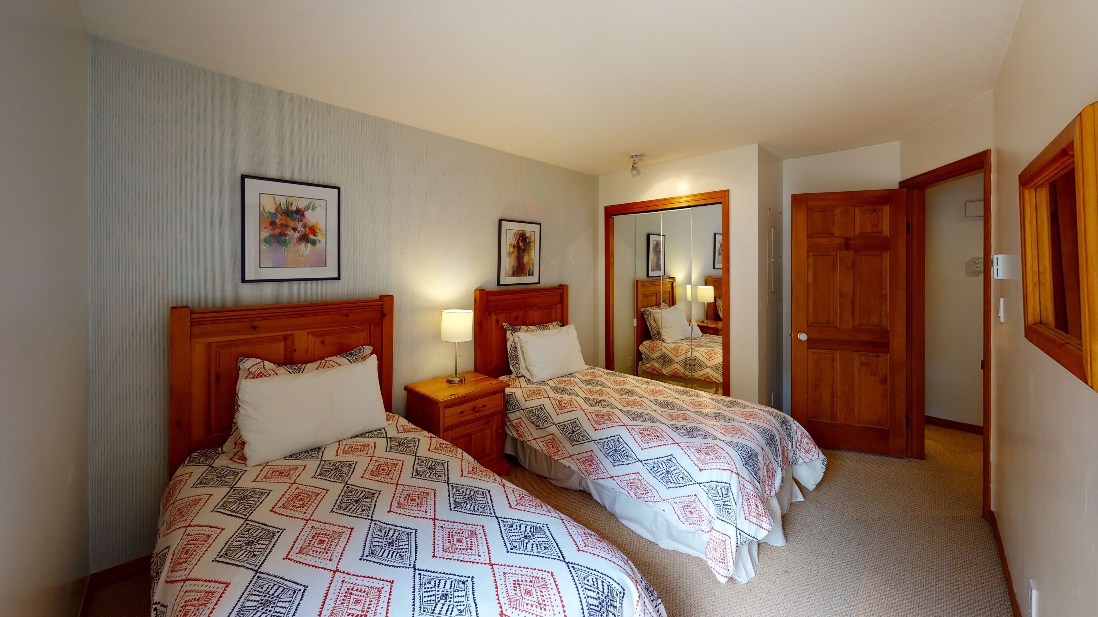 Una habitación de hotel con dos camas, cada una con ropa de cama estampada, una mesita de noche con lámpara y un espejo que refleja la habitación.