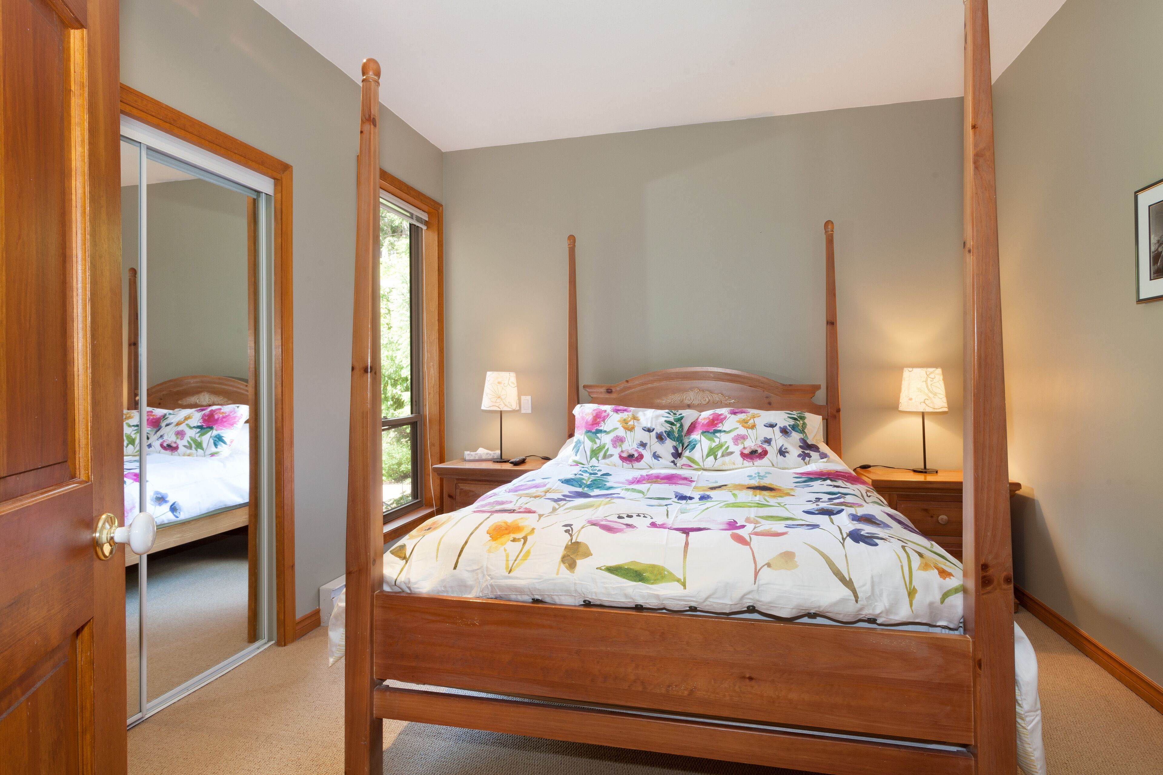 A four-poster bed with floral bedding, wooden headboard, bedside lamps, and a mirror on the door.