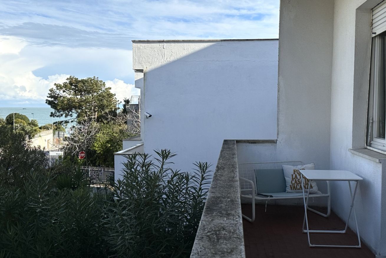 A balcony with a white wall, a small table, a bench, and a view of the sea.