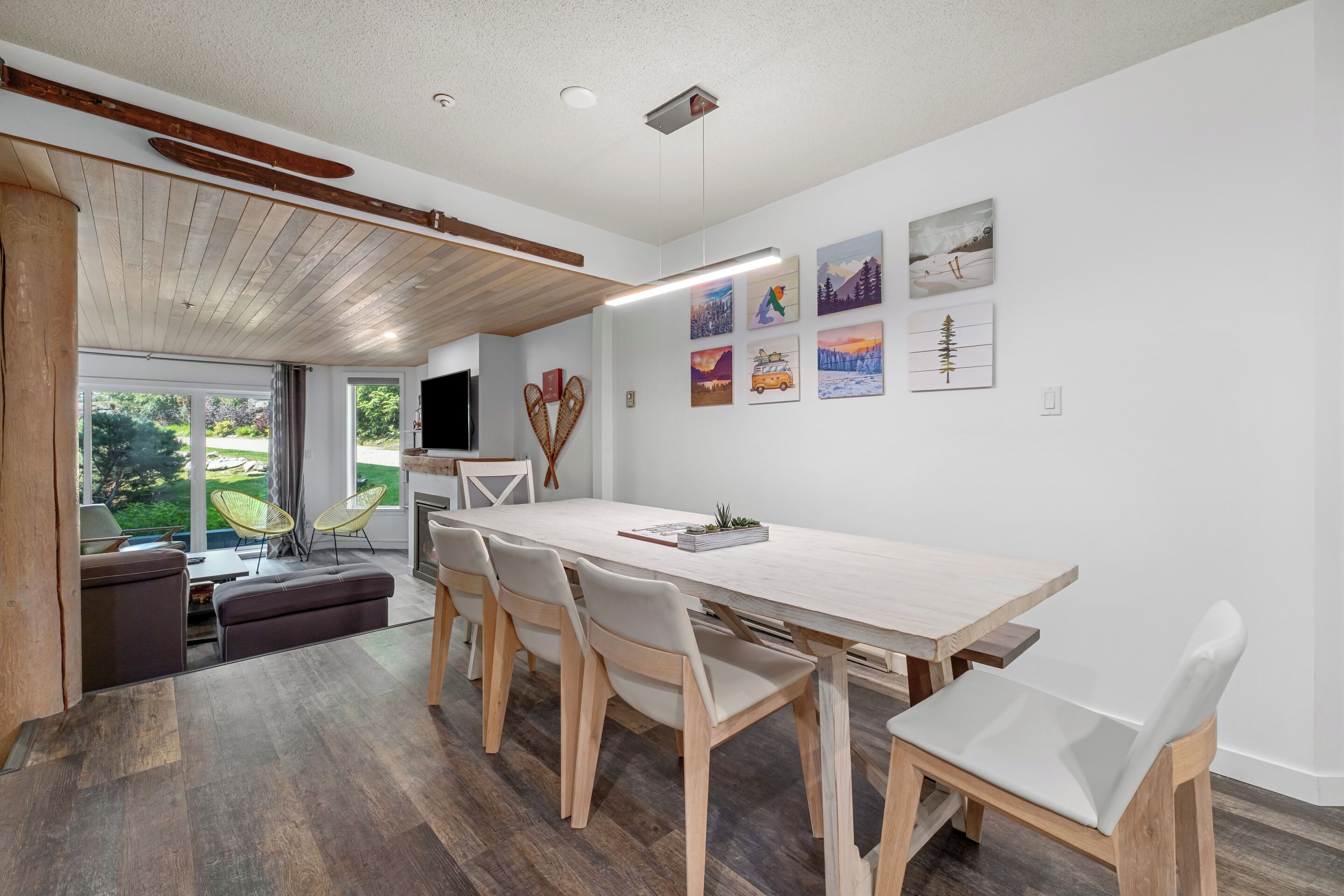 A modern dining area with a long wooden table, white chairs, and a view of the outdoors through large windows.
