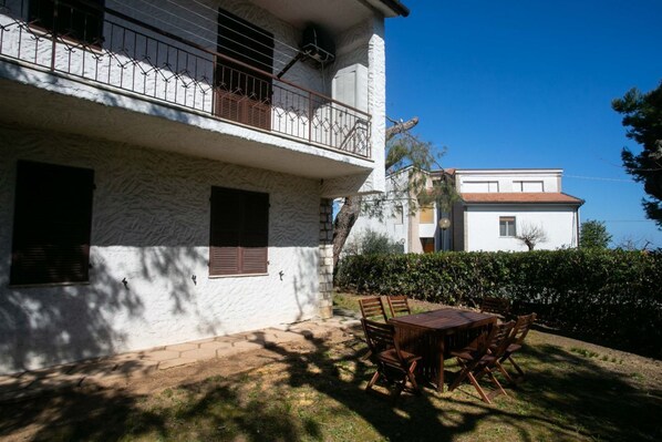 A two-story house with a balcony, a patio with a wooden table and chairs, and a hedge in the background.
