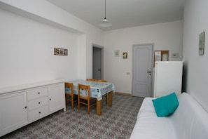 A small, clean room with a white sofa, a dining table with chairs, a white cabinet, and a patterned floor.