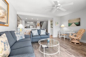 A modern living room with a grey sofa, a glass coffee table, a wicker chair, and a ceiling fan.