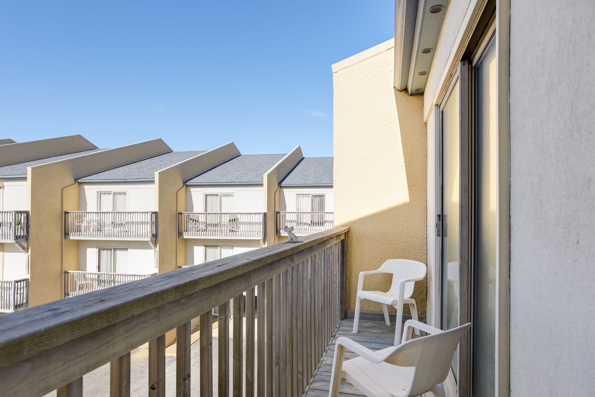 A balcony with wooden railings, white plastic chairs, and a view of adjacent buildings.