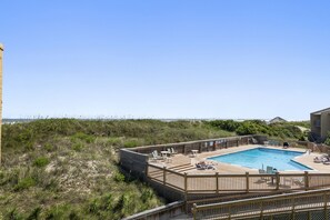 A wooden deck with a pool, lounge chairs, and a view of the beach.