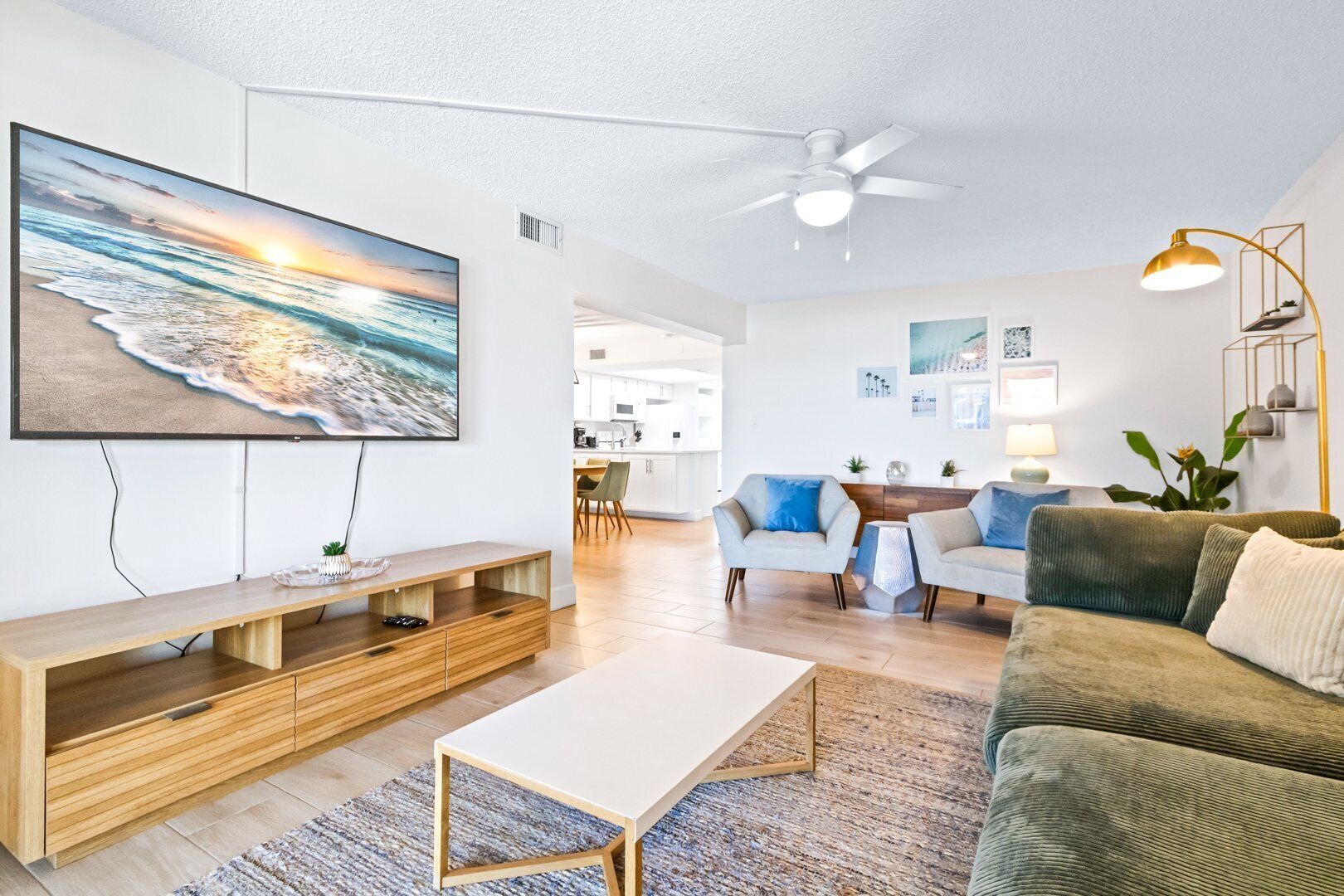 A modern living room with a flat-screen TV, a ceiling fan, and a dining area with a kitchen in the background.