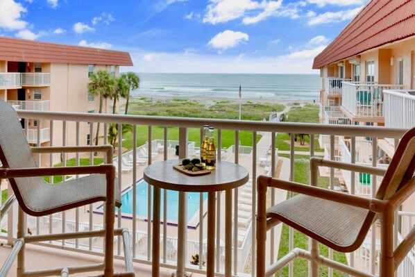 A balcony with two wicker chairs and a small table, overlooking a pool and a beach.