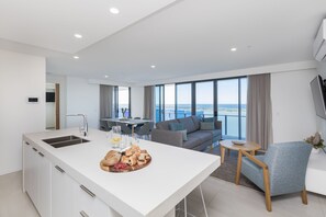 A modern kitchen with a central island, dining area with a table and chairs, a grey sofa, and a view of the sea. - Sky Broadwater Apartments (Gold Coast)