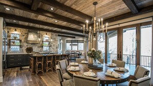 Un comedor amplio con una mesa de madera dispuesta para seis personas, rodeada de sillas. Un candelabro cuelga encima, y la habitación cuenta con vigas de madera a la vista en el techo.