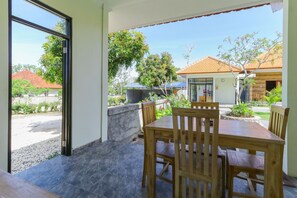 A patio with a wooden table and chairs, a stone wall, and a view of a residential area with houses and trees. - Manik Homestay Uluwatu by Zuzu (Pecatu)