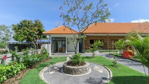 A house with a tiled roof, a garden with a circular stone planter, and a small tree with pink flowers. - Manik Homestay Uluwatu by Zuzu (Pecatu)