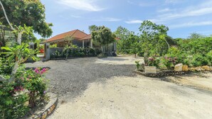 A house with a gravel driveway, surrounded by lush greenery and a clear sky. - Manik Homestay Uluwatu by Zuzu (Pecatu)
