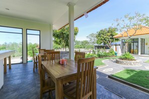 Un área de comedor al aire libre con techo, con una mesa y sillas de madera, una jardinera de piedra y vistas a la vegetación y edificios circundantes.