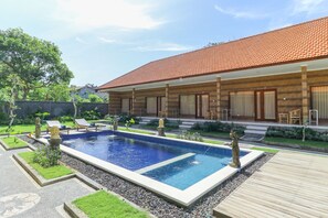 A poolside area with a rectangular pool, wooden deck, and a building with a tiled roof. - Manik Homestay Uluwatu by Zuzu (Pecatu)