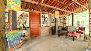 A rustic interior with wooden beams, artwork on the walls, a wooden door, a sofa, a table with a red cloth, and a chair. - Segajih Central Stay and Education (Kokap)