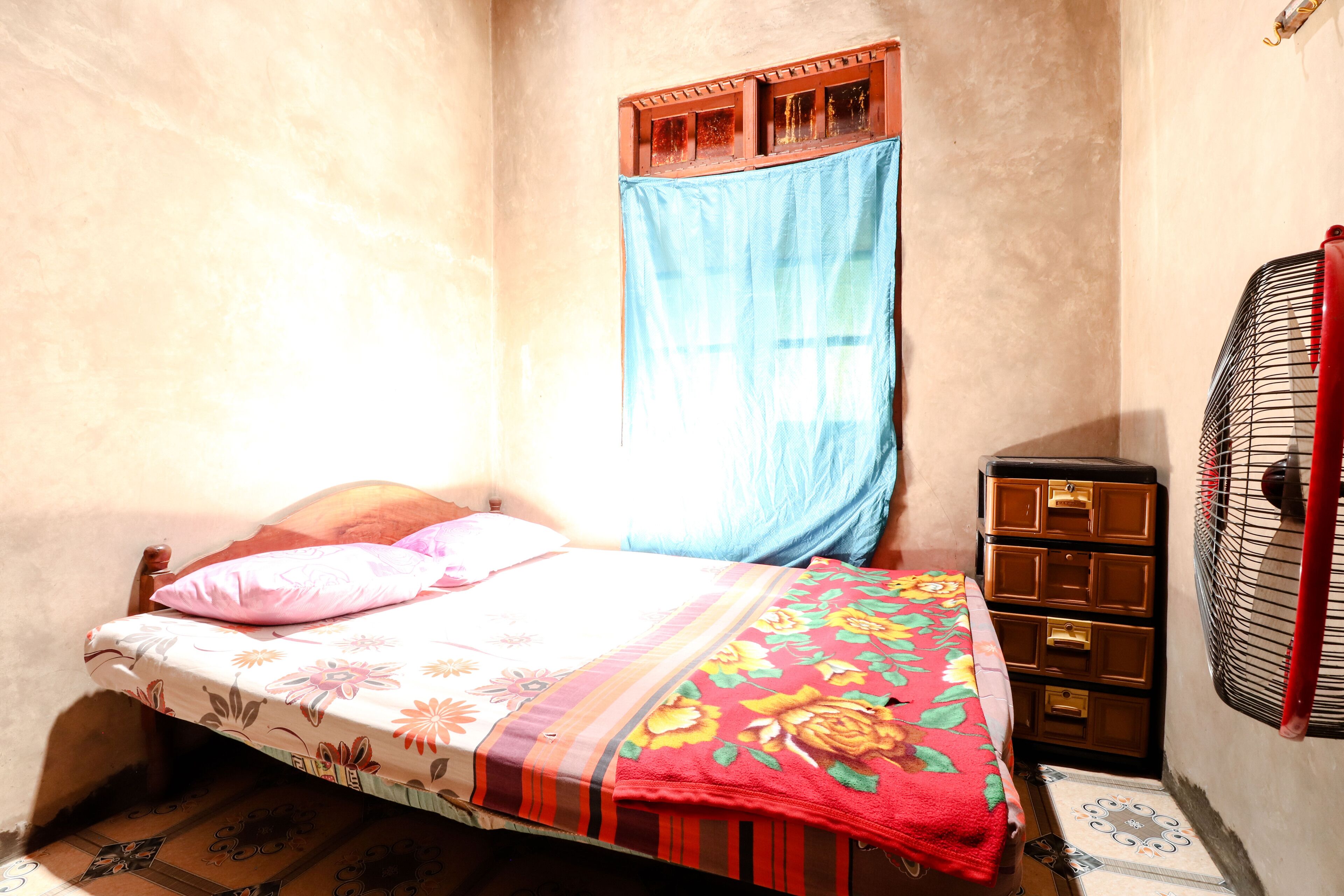 Una habitación pequeña y tenuemente iluminada, con una cama individual, un tocador de madera y una ventana con cortina azul.