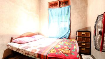 Una habitación pequeña y tenuemente iluminada, con una cama individual, un tocador de madera y una ventana con cortina azul.