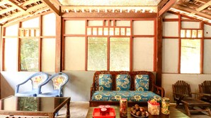 A traditional room with a wooden sofa, blue chairs, a wooden table with food items, and large windows with wooden frames.