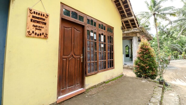 Um edifício amarelo com uma grande porta de madeira e janelas, uma placa indicando um lar de hóspedes e uma pequena árvore em primeiro plano.