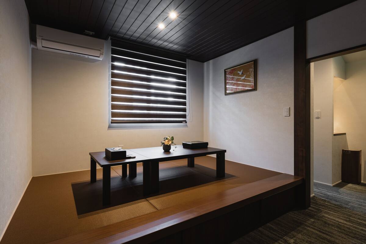 A room with a tatami mat floor, a low wooden table with a vase, and a window with blinds.