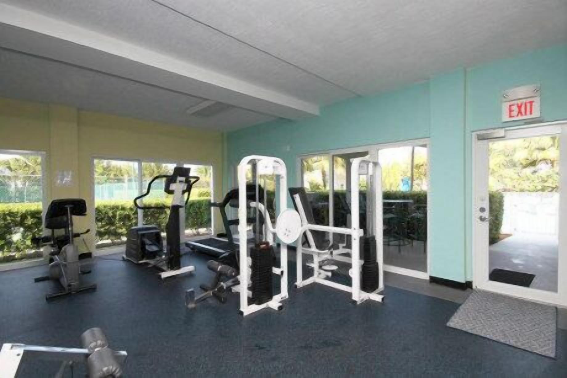A gym with various exercise machines, a door marked 'EXIT', and a view of greenery outside.