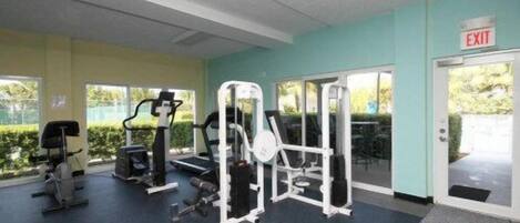 A gym with various exercise machines, a door marked 'EXIT', and a view of greenery outside.