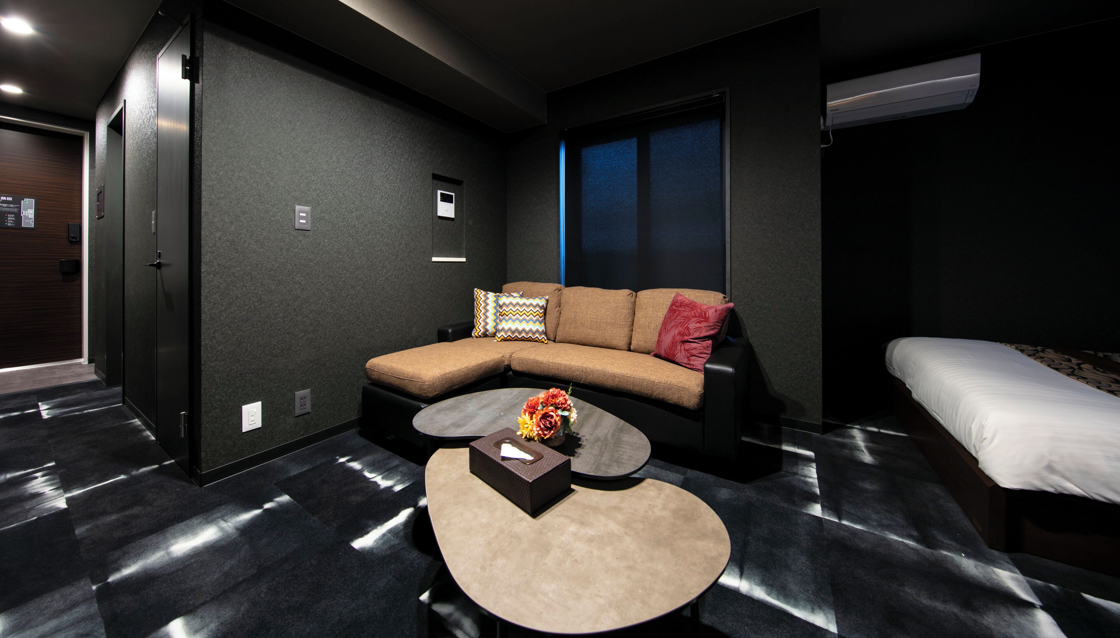 A modern hotel room with a sectional sofa, a small round coffee table with flowers, a bed, and a large window.