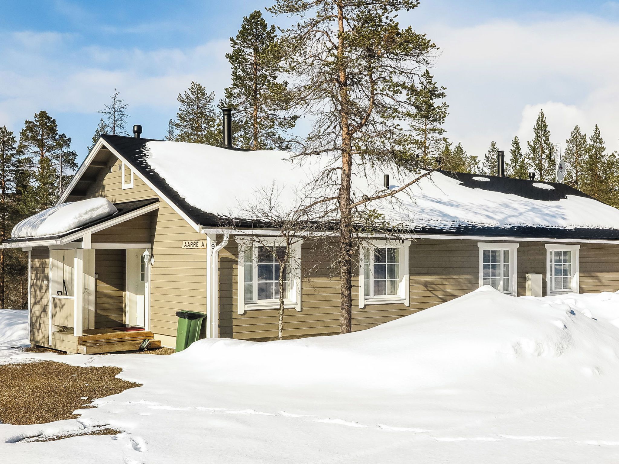 A single-story wooden cabin with a snow-covered roof, a front porch, and a visible chimney.