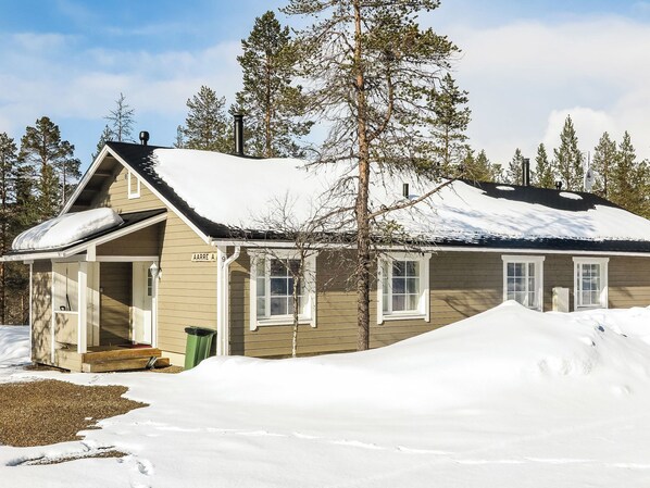 A single-story wooden cabin with a snow-covered roof, a front porch, and a visible chimney.