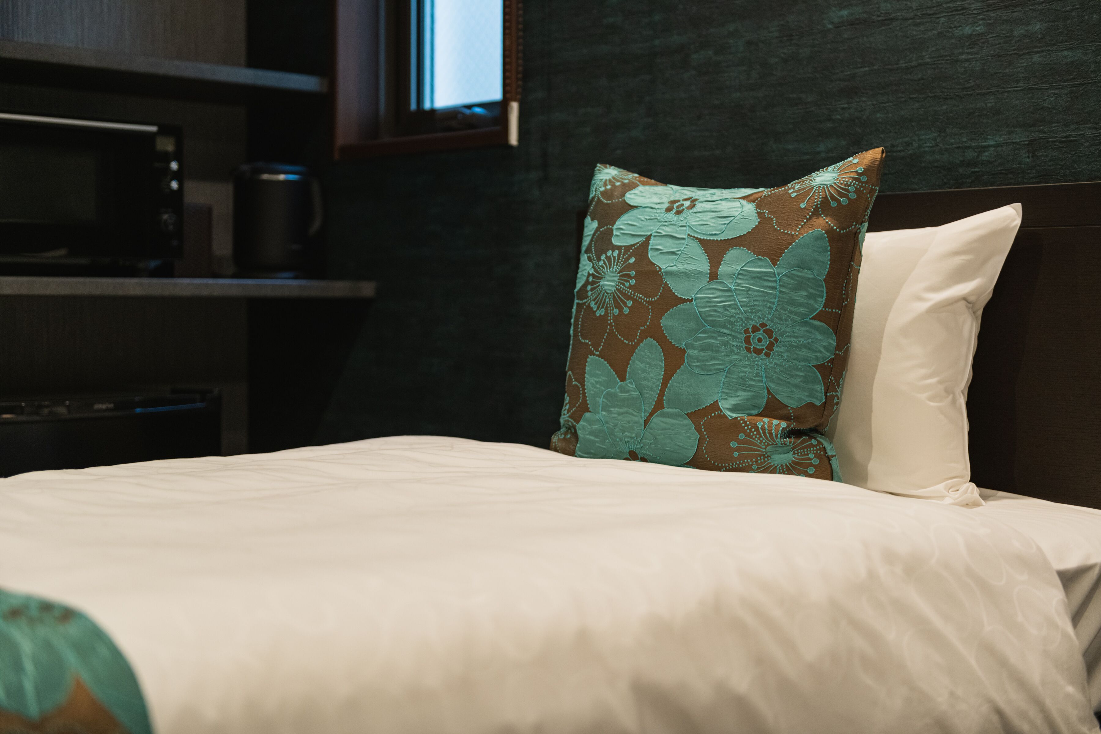 Quarto de hotel com cama, travesseiro estampado com flores, mesa de cabeceira e cafeteira, e cabeceira escura.