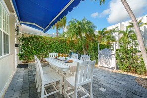 Une terrasse extérieure avec une table et des chaises blanches, un auvent bleu et une vue sur des palmiers et des bâtiments en arrière-plan.