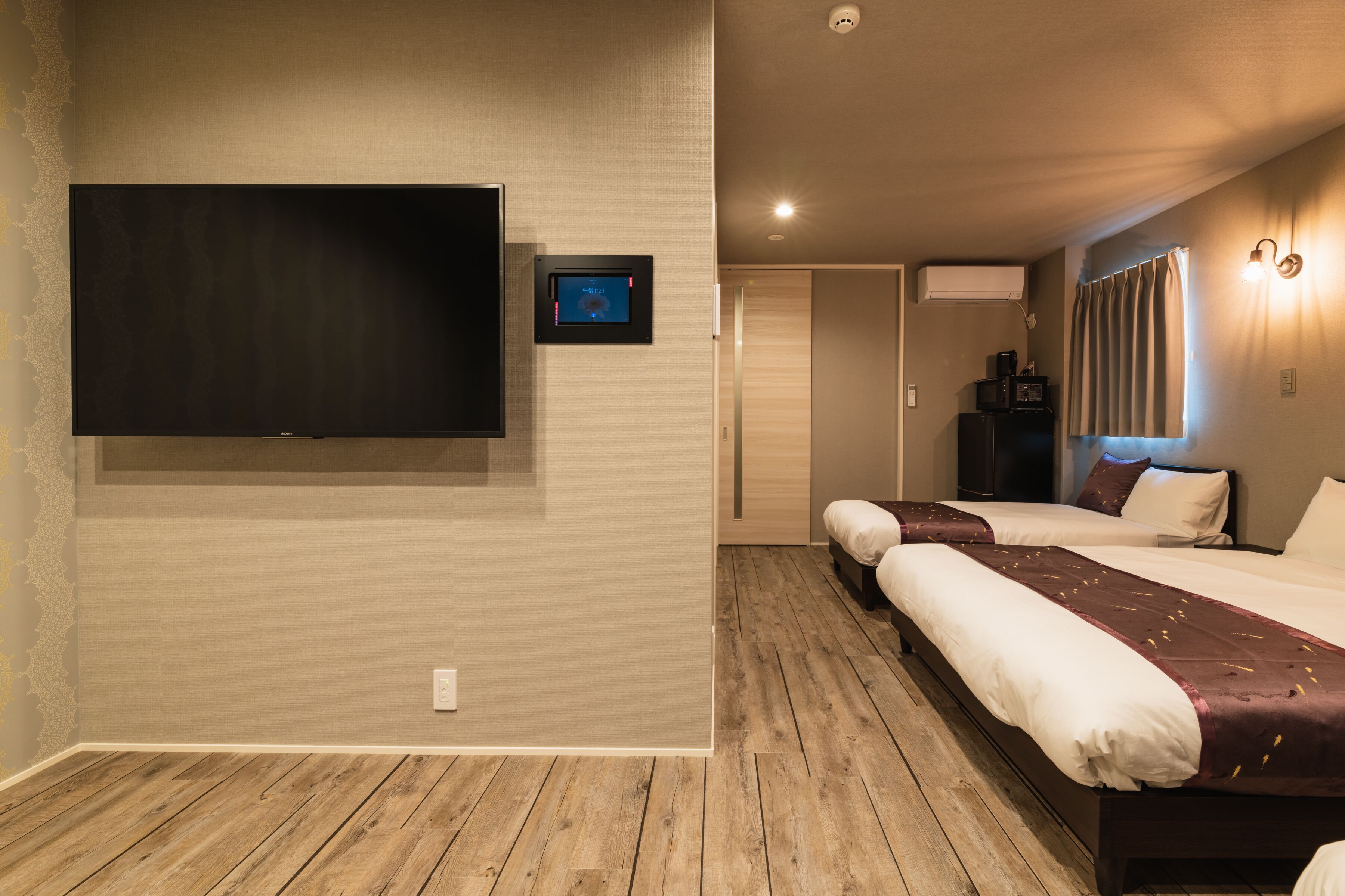 A hotel room with a large flat-screen TV mounted on the wall, a wooden floor, two beds with white bedding, a small bedside table with a lamp, and an air conditioning unit.