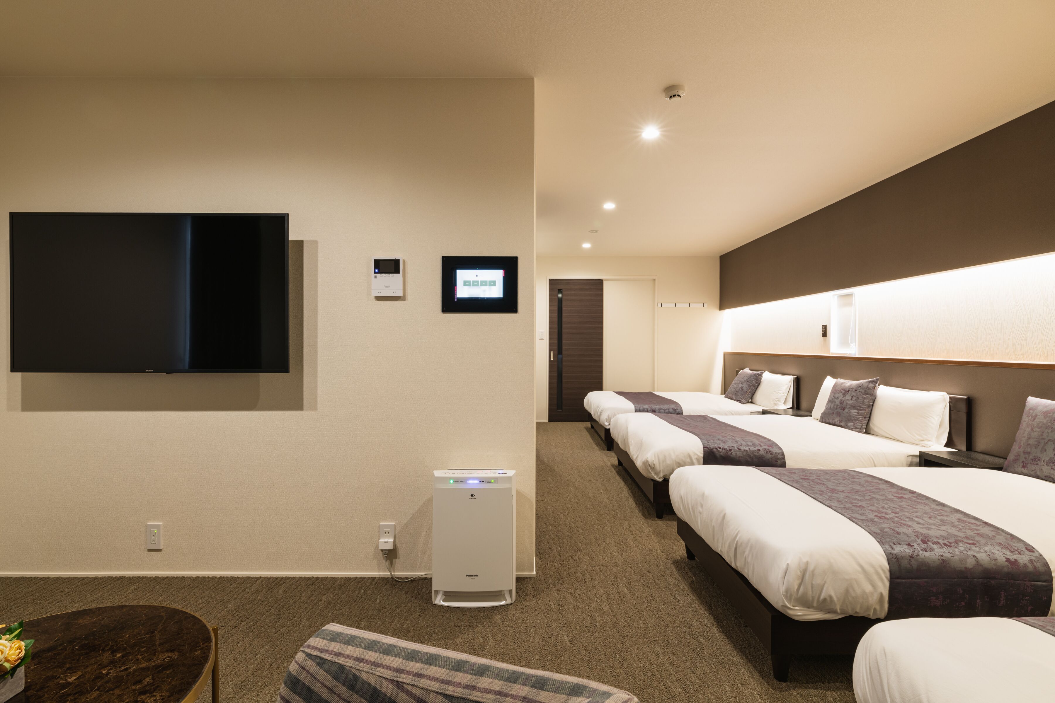 Una habitación de hotel con una televisión de pantalla plana grande montada en la pared, un minibar, una cama con cabecera y una mesita de noche.