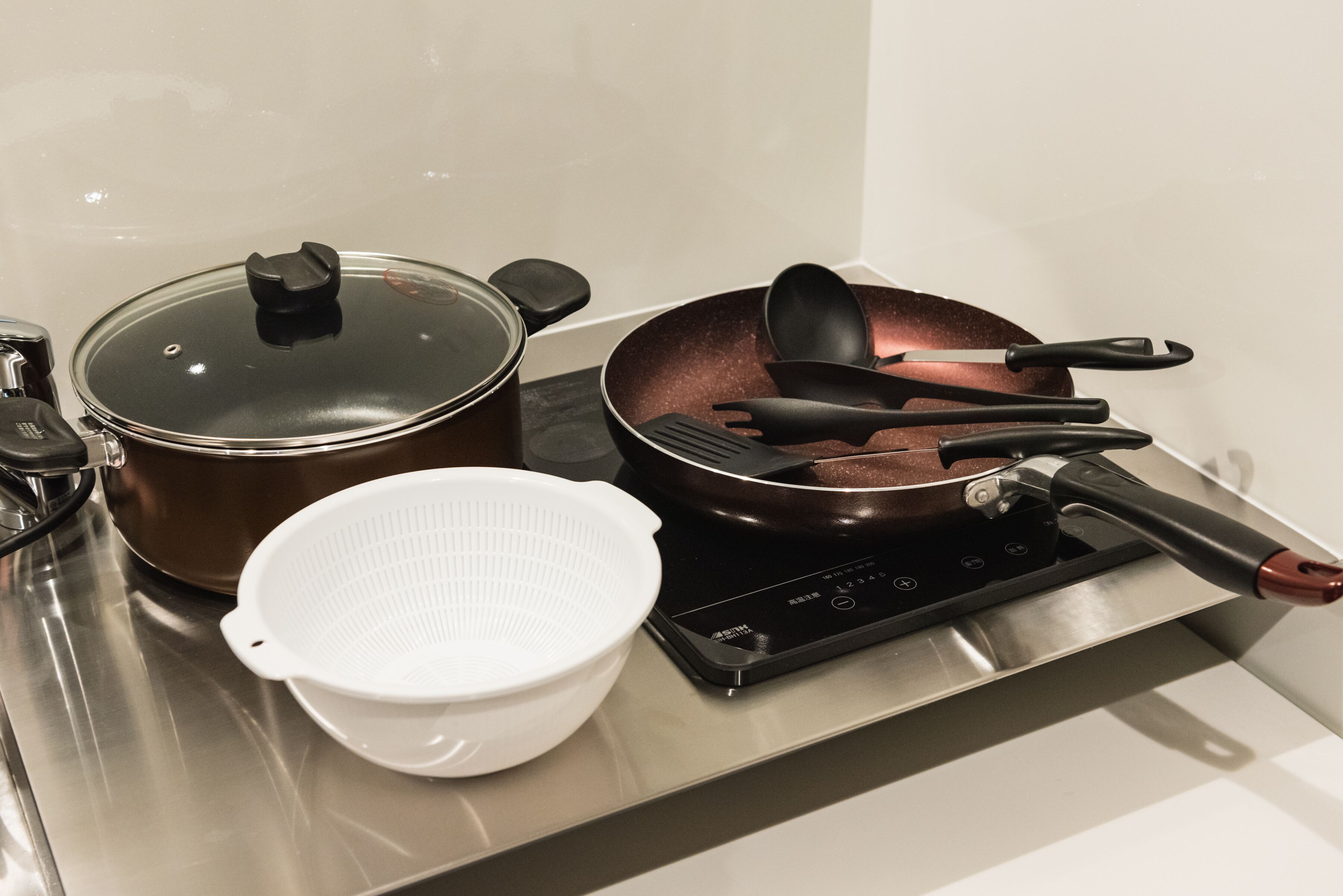A kitchen with a stainless steel stove, a brown pot with a lid, a white colander, and a frying pan with black utensils.