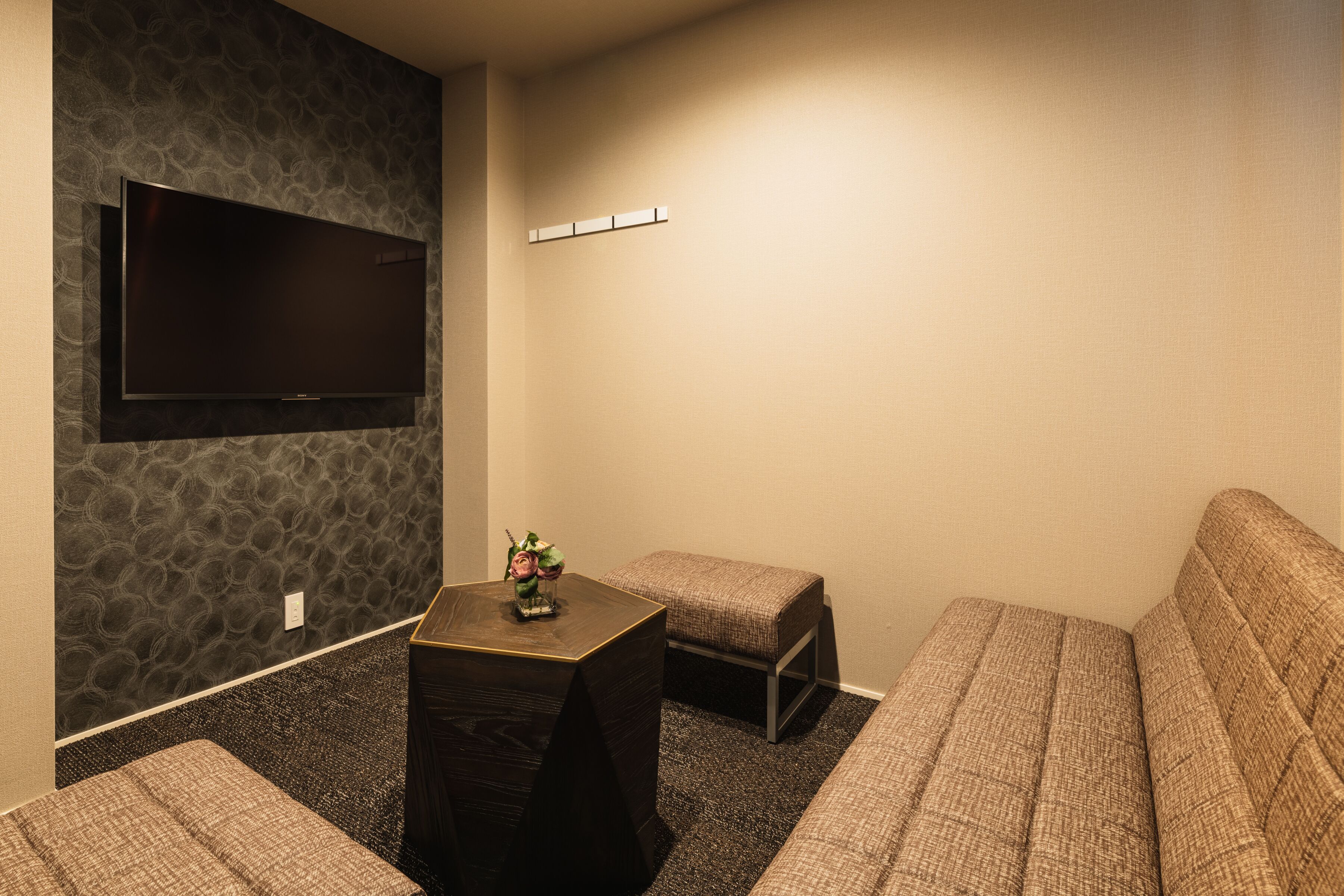A room with a flat-screen TV mounted on a textured wall, a small table with a decorative item, and two upholstered seating areas.