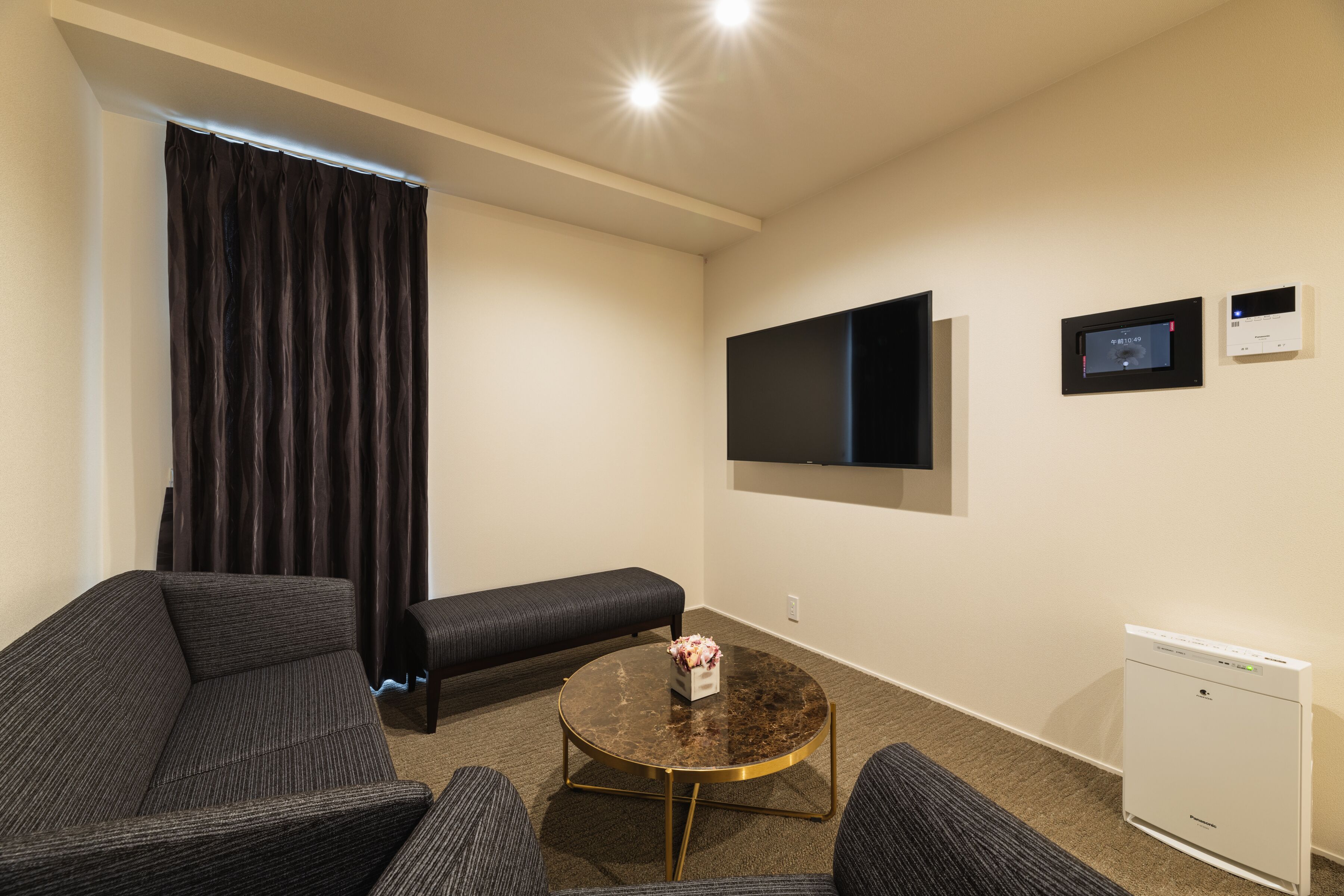 Moderni hotellihuone, jossa on sohva, pieni pyöreä pöytä maljakolla, penkki, seinälle asennettu televisio ja pieni valkoinen kodinkone.