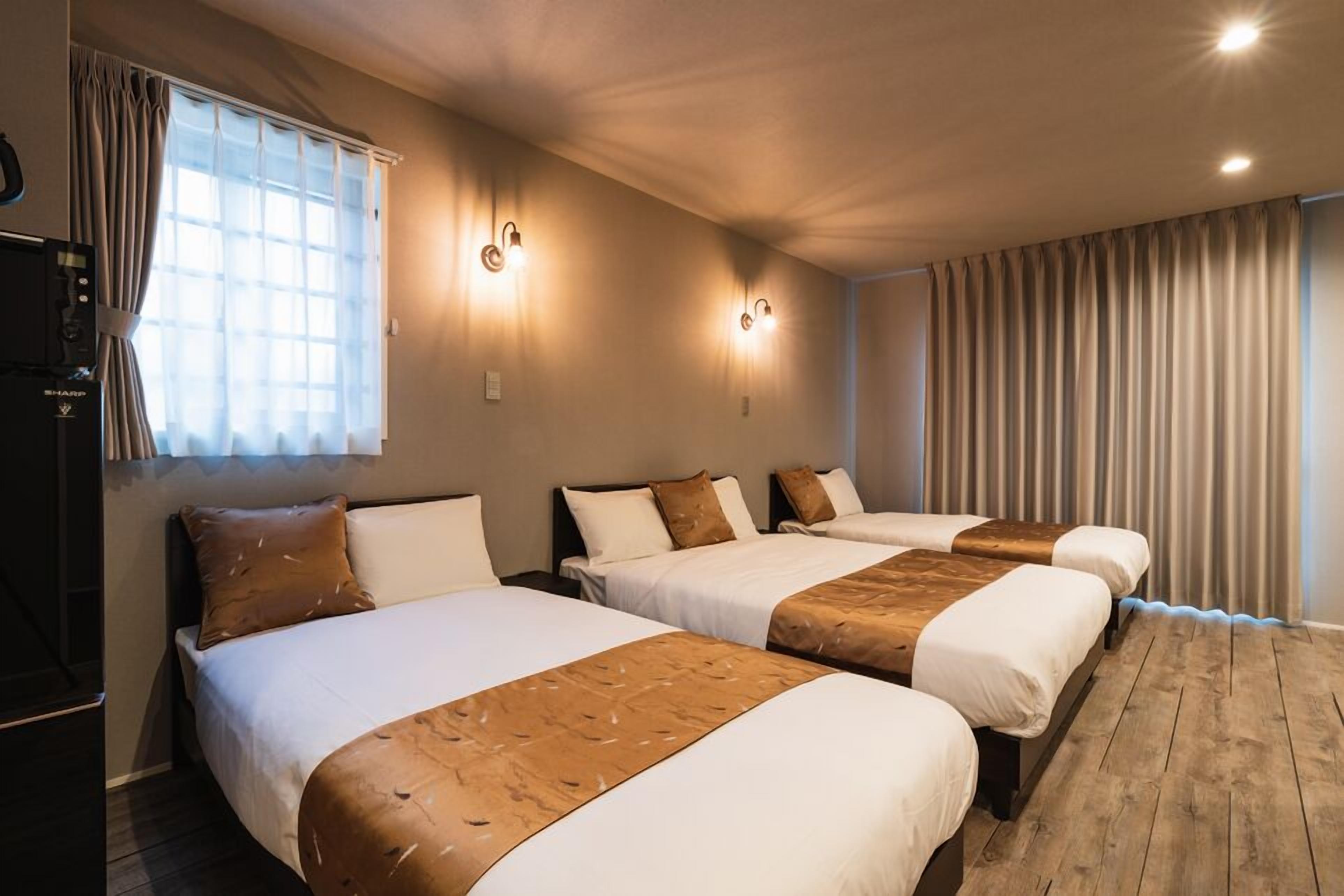 A hotel room with three beds, a large window with curtains, wall-mounted lights, and a wooden floor.