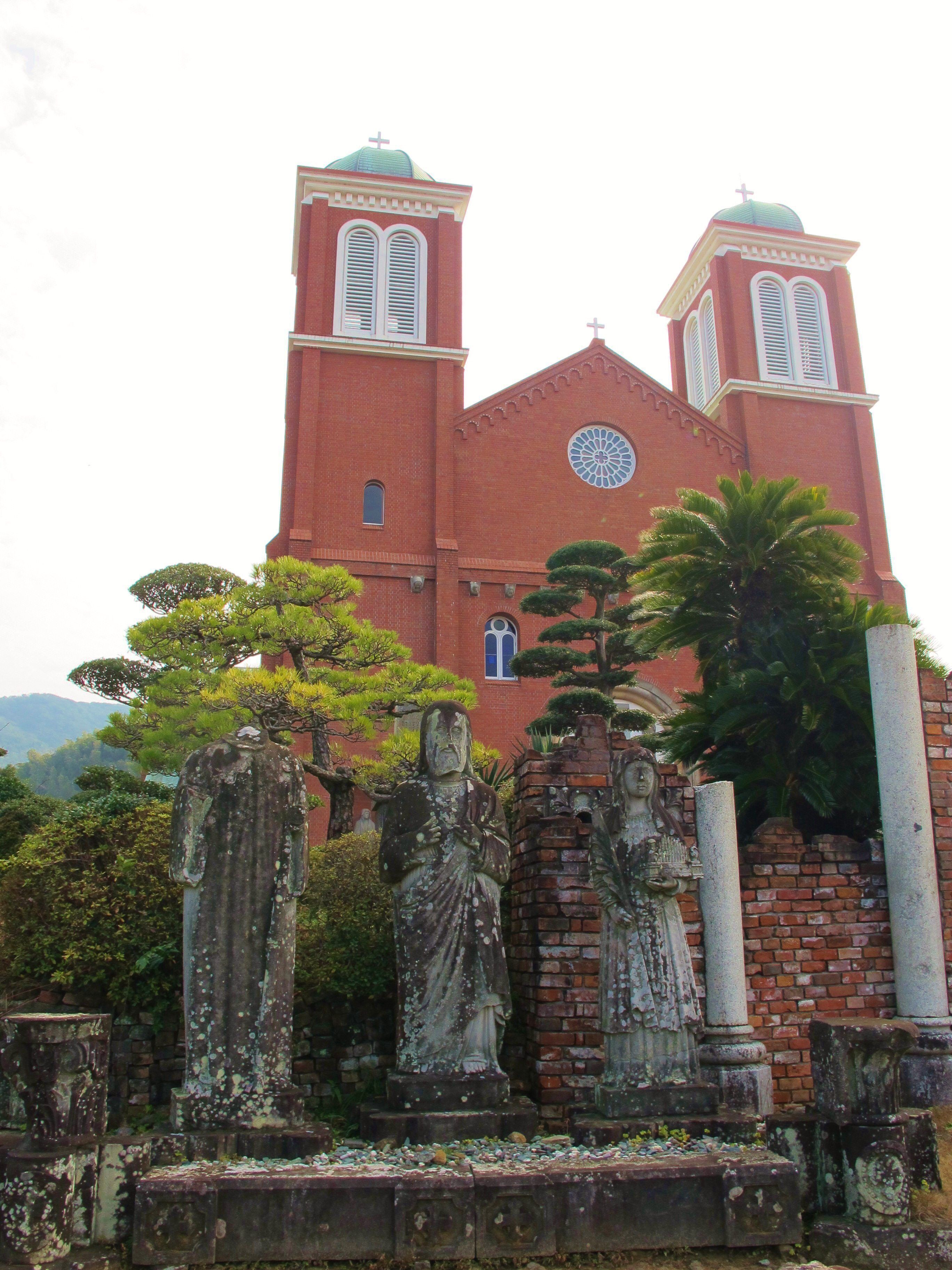 A red brick church with two bell towers and a large circular window. In front of the church, there are stone statues and a palm tree.