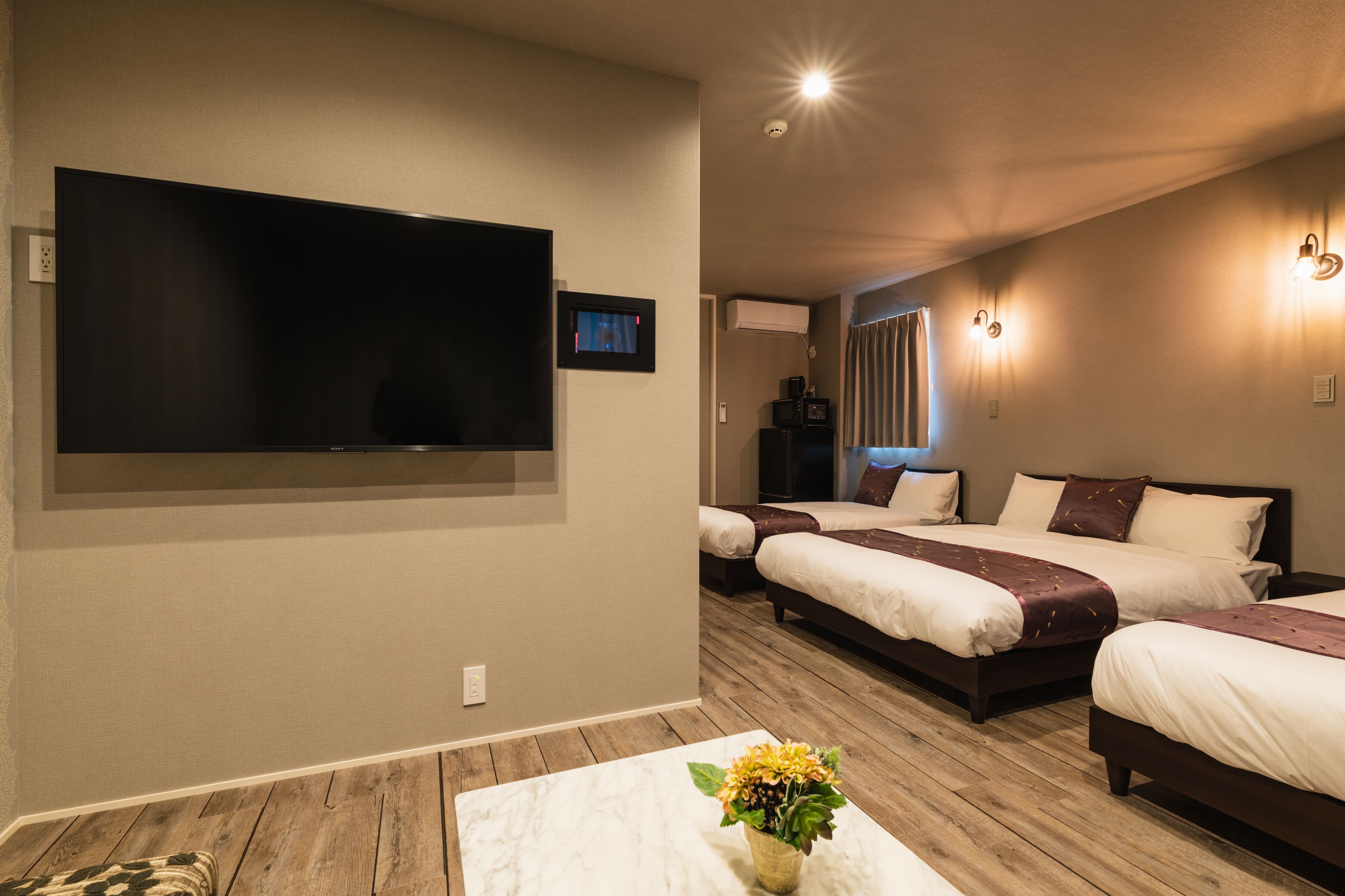 A hotel room with a large flat-screen TV mounted on the wall, two beds with white bedding and brown headboards, a small table with a vase of flowers, and a ceiling light fixture.