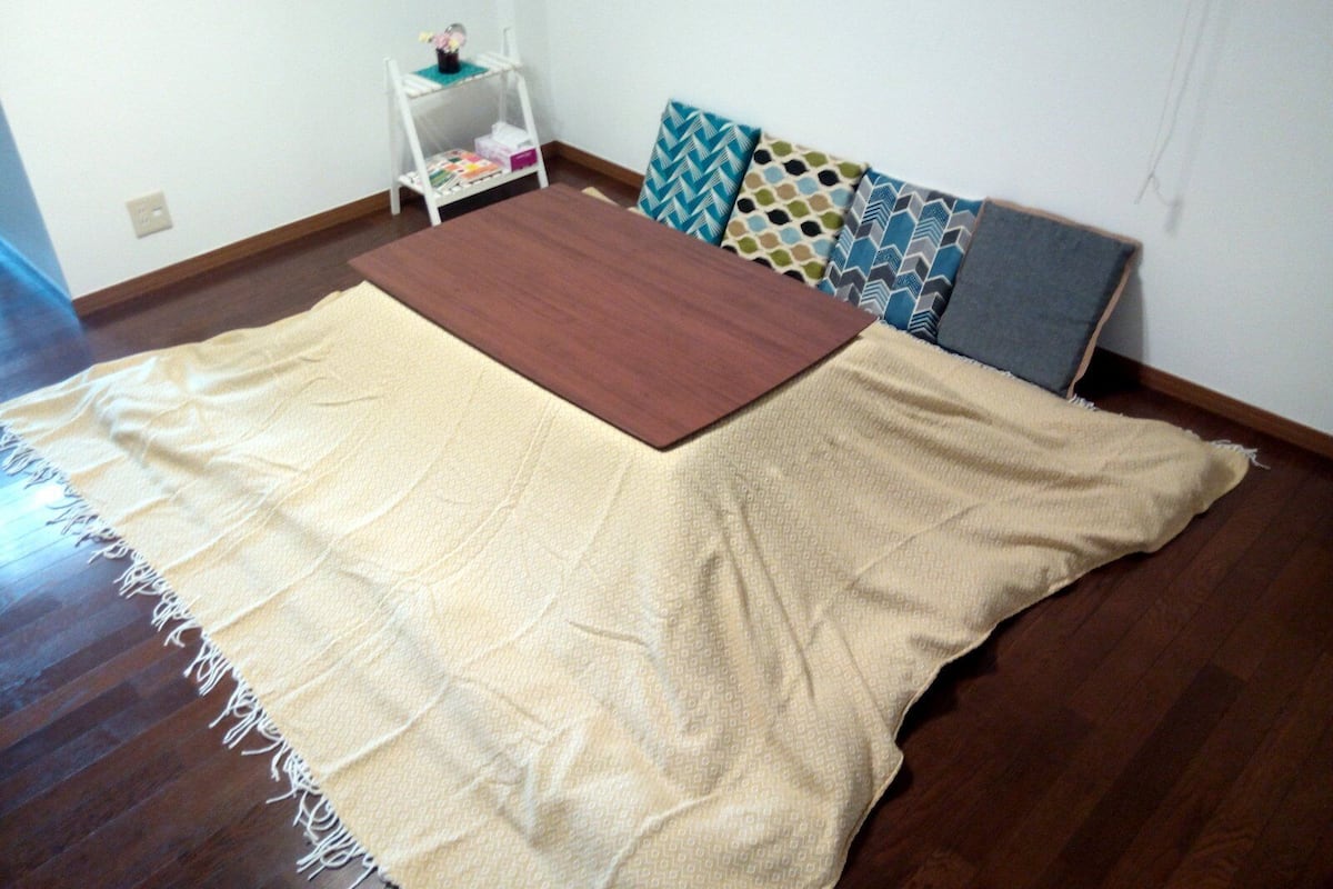 Una habitación con una cama grande cubierta con una manta color beige, piso de madera y una mesita de noche blanca con una lámpara pequeña y libros.
