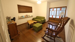 A living room with a wooden floor, a green sofa, a wooden rocking chair, a wooden coffee table, a wooden side table, a wooden cabinet, a wooden bookshelf, and a window with blinds. - Sumica Apartments (Nikko)