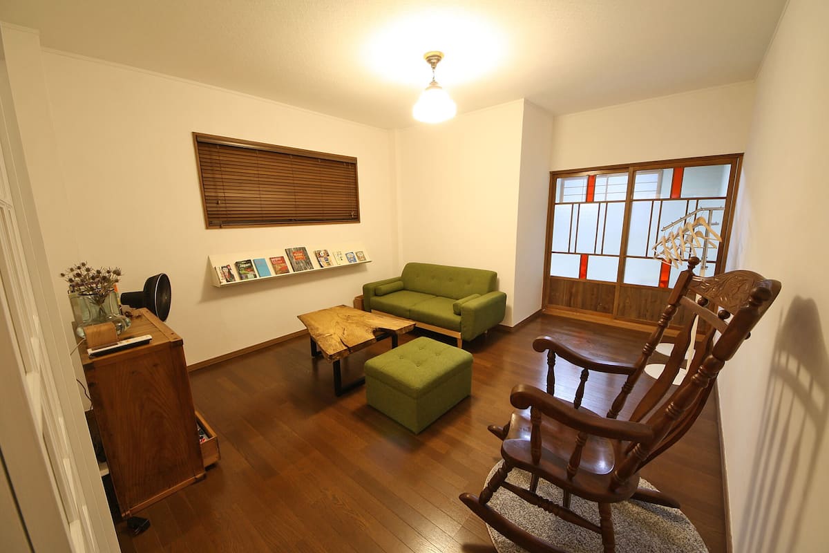 Un salón con piso de madera, un sofá verde, una mecedora de madera, una mesa de centro de madera, una mesita auxiliar de madera, un aparador de madera y una estantería de madera, además de una ventana con persianas.