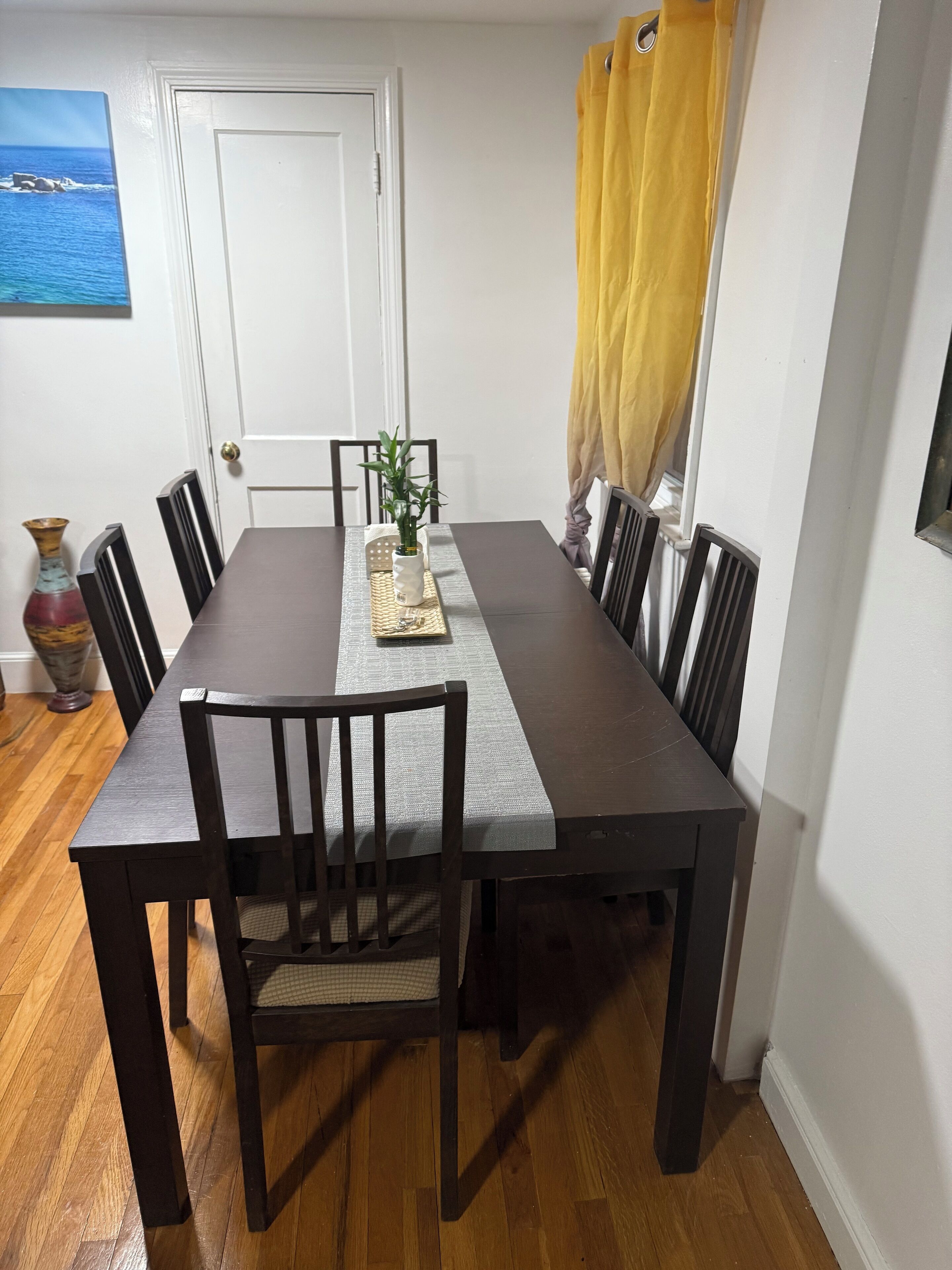 A dining room with a rectangular table, six chairs, a vase, and a painting on the wall.