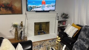 Una sala de estar con chimenea, un televisor mostrando una escena de playa, un sofá de cuero negro y una mesa de centro.