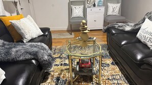 Una sala de estar con un sofá de cuero negro, una mesa de centro de vidrio y un juego de Monopoly, además de una alfombra estampada.