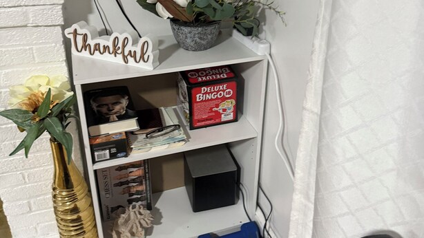 Una esquina de habitación con pared de ladrillo blanco, repisa con un letrero que dice 'thankful', planta en maceta, juego de ajedrez, caja de BINGO y una pesa rusa azul.