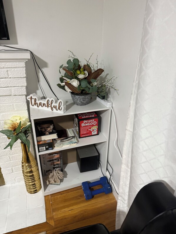 A corner of a room with a white brick wall, a shelf with a 'thankful' sign, a potted plant, a chess set, a BINGO box, and a blue dumbbell.